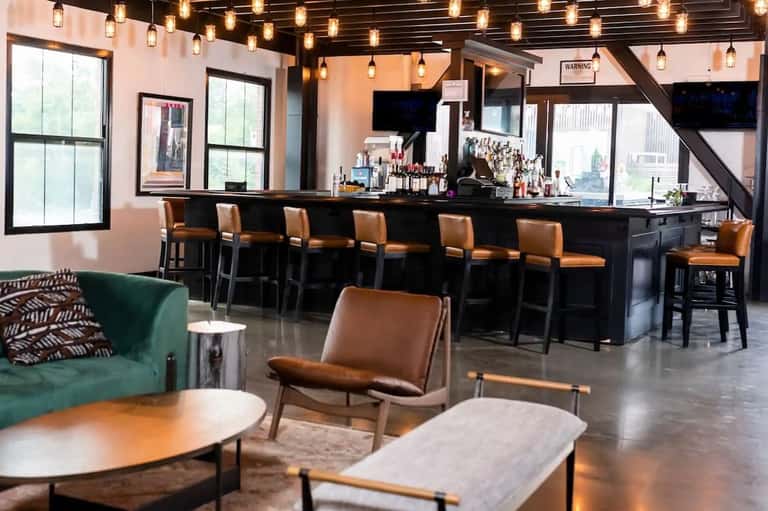 Modern bar interior with tan leather seating, dark bar counter, hanging lights, and large windows