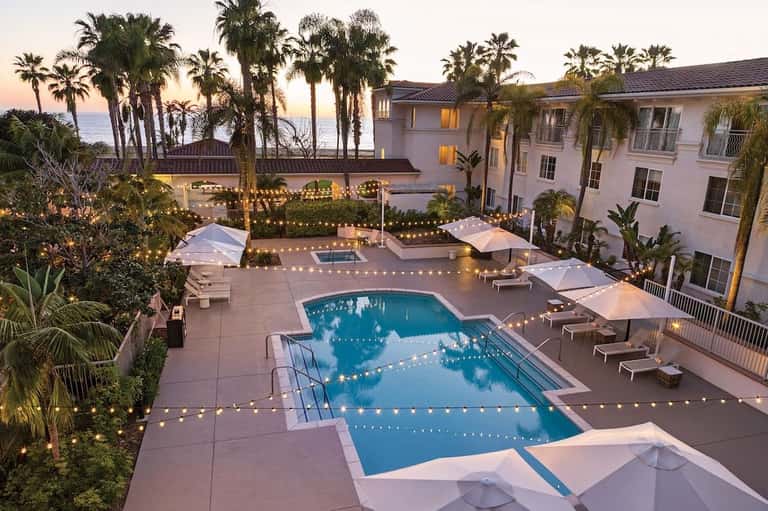 Aerial view of resort pools at dusk with string lights, umbrellas, and ocean view