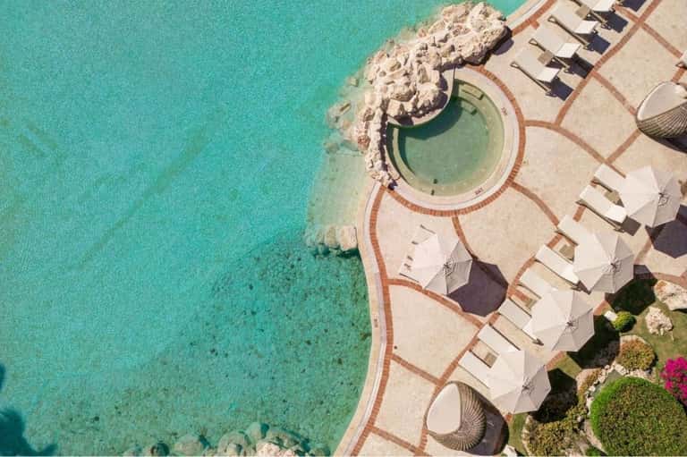 Circular pool with turquoise water overlooking the ocean, bordered by white umbrellas and loungers