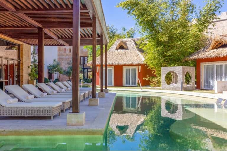 Modern spa courtyard with lounge chairs, pergola, reflecting pool, and thatched-roof treatment rooms