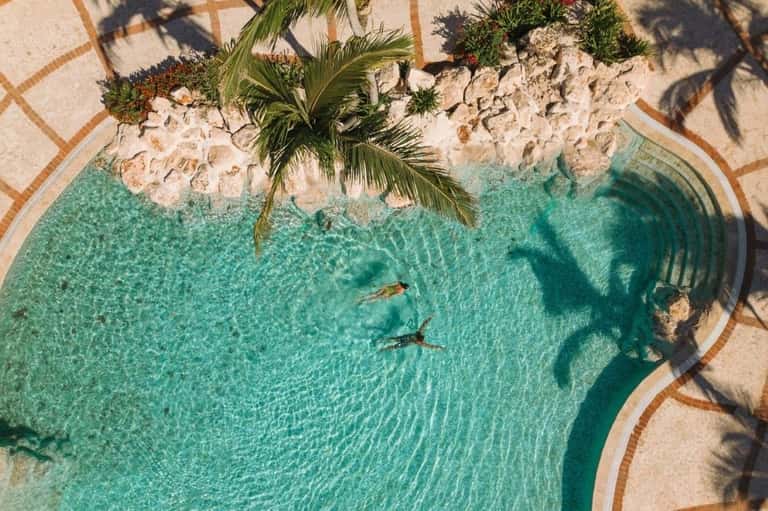 Aerial view of turquoise natural pool with swimmers and sandy beach with palm tree shadows