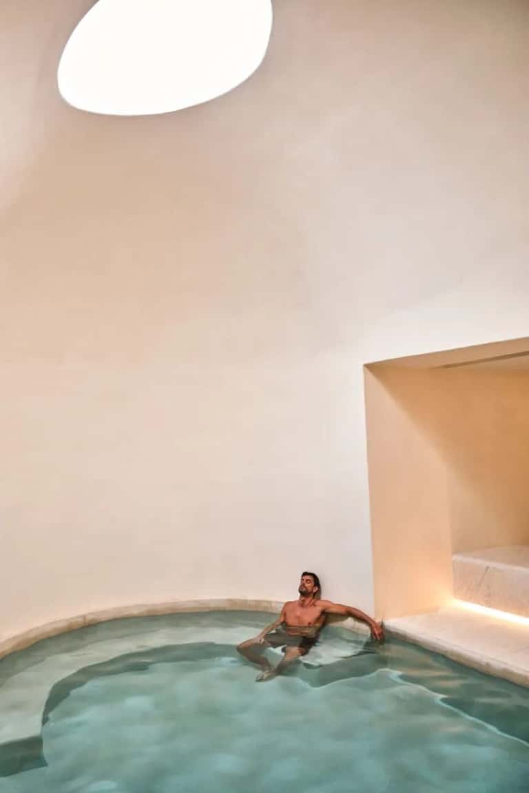 Person bathing alone in minimalist circular pool with white walls and natural skylight