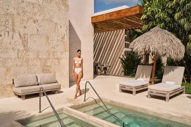 Woman in white bikini standing by geometric thermal pool with stone walls and thatched umbrellas