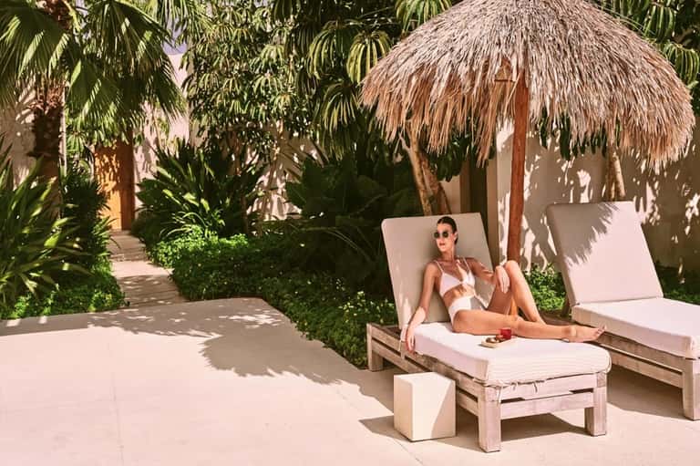 Woman in white bikini lounging under a thatched umbrella by a wooden sun chair in a tropical garden courtyard