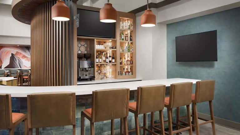 Modern hotel bar with copper pendant lights, white counter, wooden chairs with orange cushions, and teal accent wall with mounted TV