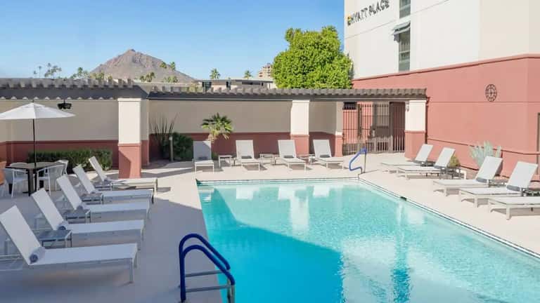 Resort rooftop pool with crystal blue water, white lounge chairs, pink building facade, and mountain views in desert setting