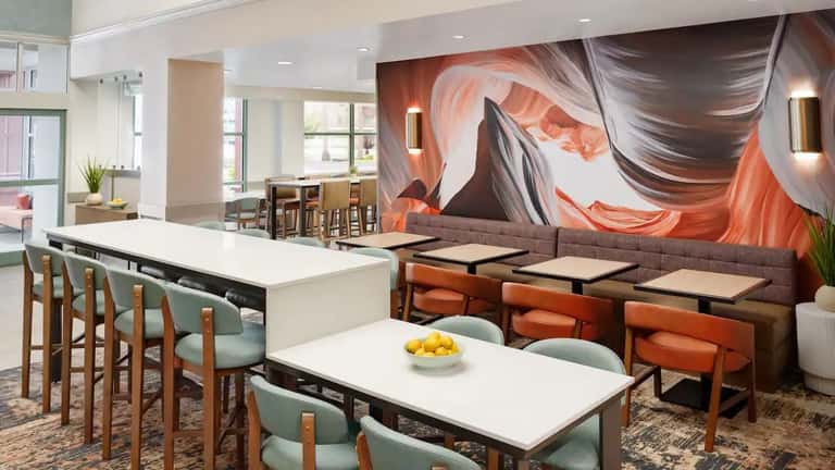 Hotel lobby lounge with white dining tables, wooden chairs, large abstract orange wall art, and floor-to-ceiling windows