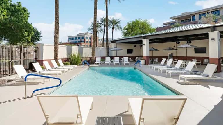 Outdoor resort pool with clear turquoise water, white loungers, palm trees, covered patio area, and modern building architecture