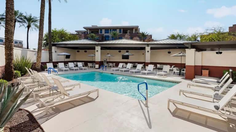 Hotel courtyard pool featuring bright turquoise water, tan loungers, palm trees, covered pavilion with fans, and terra cotta accent walls