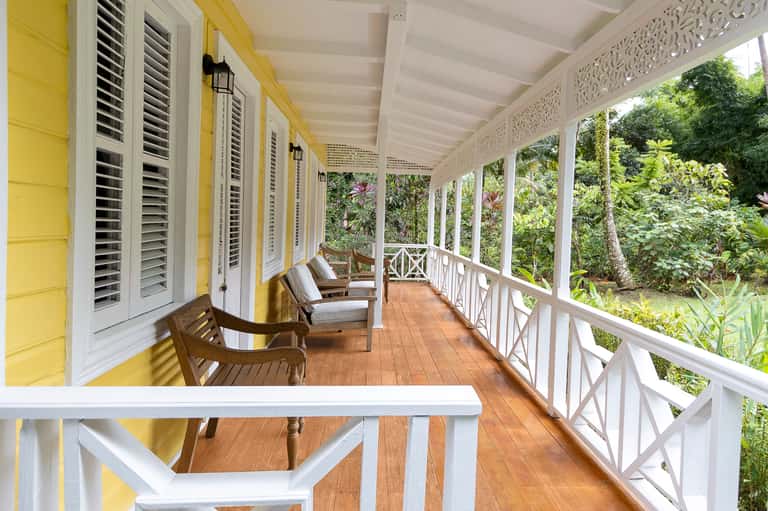 Long yellow and white covered porch with wooden chairs, shuttered doors, and views of tropical gardens
