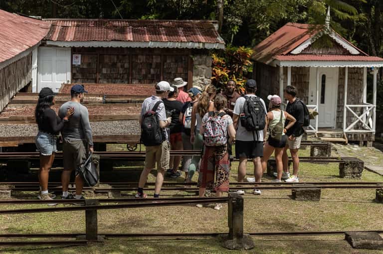 Group of tourists gathered at a colonial-era village site with corrugated metal-roof buildings