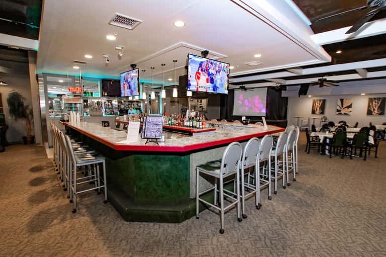 Modern bar lounge with curved counter, red and green accents, white bar seating, flat-screen TV, and ambient lighting