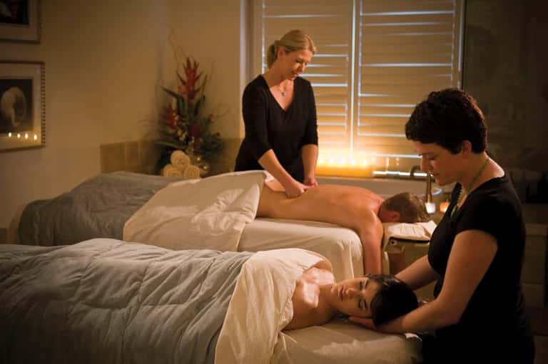 Two massage therapists performing treatments on clients lying on massage beds in a spa room with warm lighting