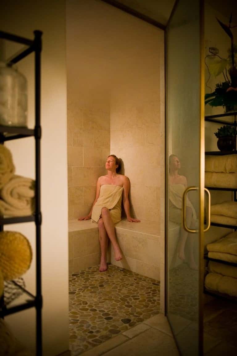 Person sitting in a steam room or sauna with tiled walls and glass enclosure door