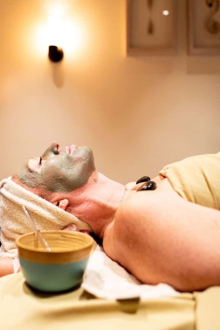 Spa therapist applying gray facial mask to relaxed male client with treatment products nearby