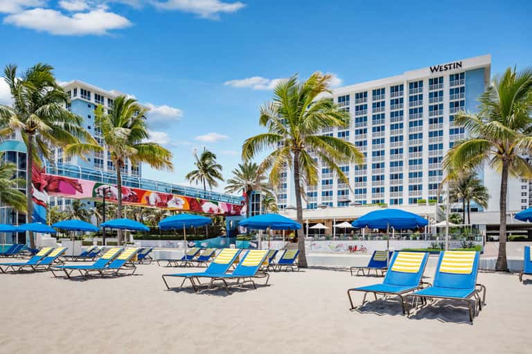Beachfront hotel with blue loungers, umbrellas, and palm trees under clear sky