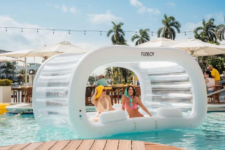 Transparent bubble lounger in resort pool with two people inside, palm trees and white umbrellas in background