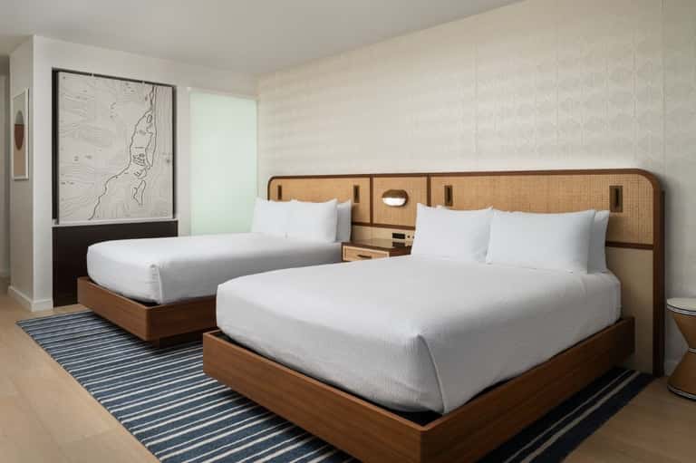 Hotel room with two double beds, rattan headboards, striped area rug, and minimal decor