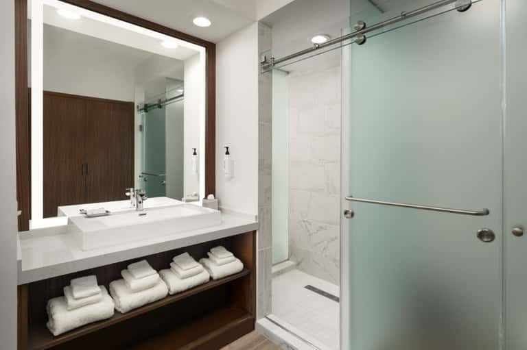 Modern hotel bathroom with illuminated vanity mirror, white sink, wooden accents, and walk-in shower