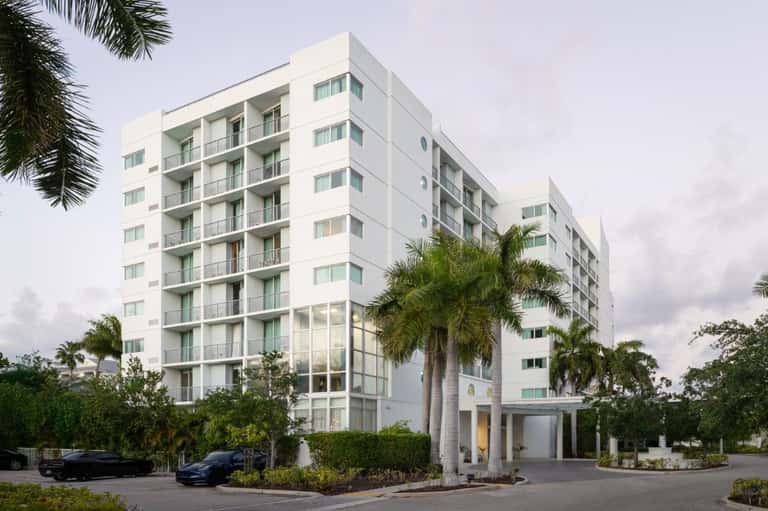 White mid-rise residential building with balconies, palm trees, and covered carport entrance