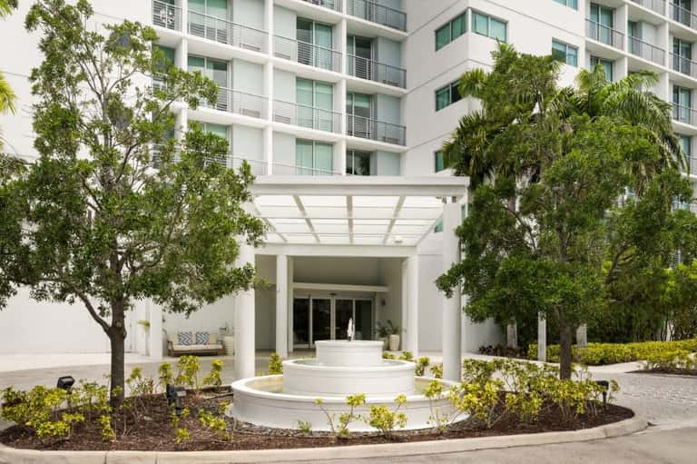 Modern hotel entrance with white columns, covered pergola, circular fountain, and mature landscaping