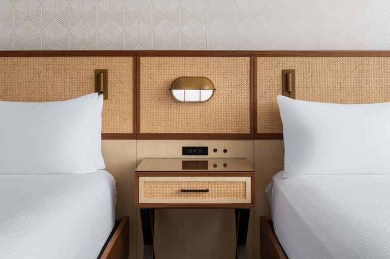 Twin hotel beds with woven rattan headboards separated by integrated wooden nightstand with drawer and round mirror