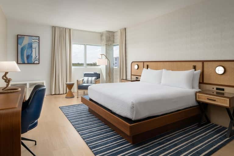 Modern hotel suite with rattan headboard bed, waterfront balcony view, white bedding, blue striped rug, desk workspace, and contemporary furnishings