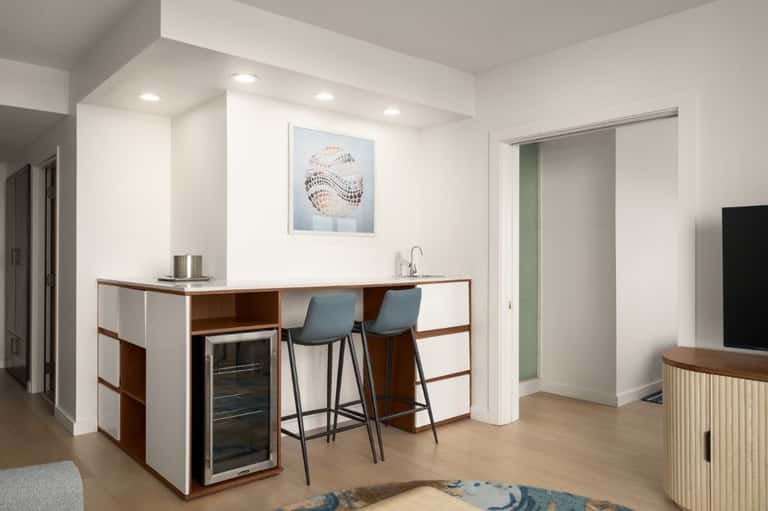 Hotel kitchenette with wooden cabinetry, white countertop, built-in wine cooler, bar seating, and blue-upholstered stools