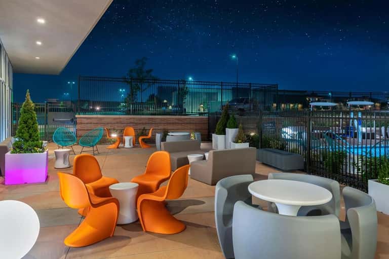 Rooftop lounge at night with orange chairs, white tables, and city skyline under starry sky