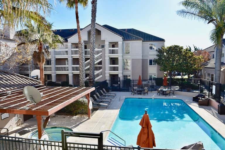 White multi-story resort building with blue swimming pool, palm trees, lounge chairs, and orange umbrellas in courtyard