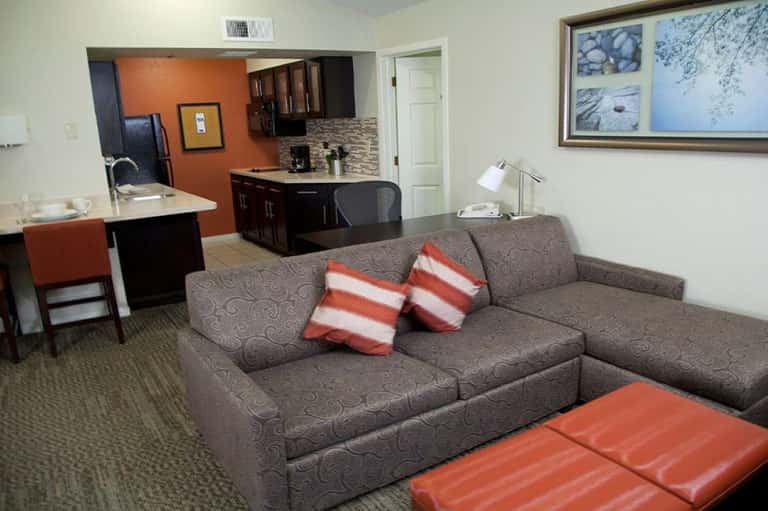 Suite living room with gray sectional sofa, orange accent wall, dark kitchen cabinetry, and striped throw pillows