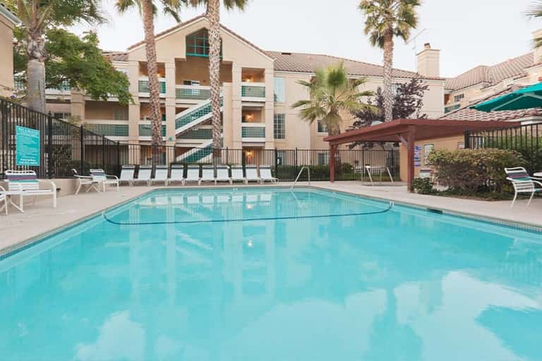 Resort outdoor pool with turquoise water, white lounge chairs, palm trees, and tan building with metal railings