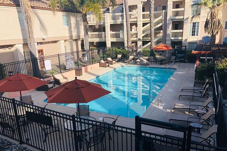Resort pool area surrounded by beige multi-story buildings, red umbrellas, lounge seating, and palm trees