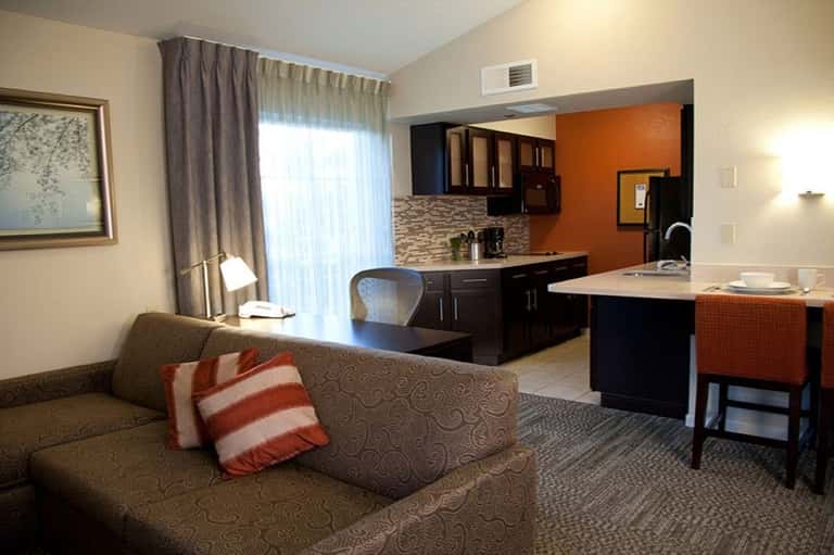 Hotel suite with living area featuring tan sofa, kitchenette with dark cabinets, orange accent wall, and large windows with curtains