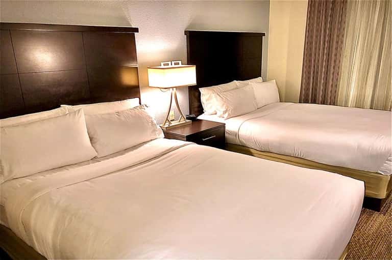 Hotel room with two queen beds, dark wood headboards, white linens, bedside lamp, and curtained windows