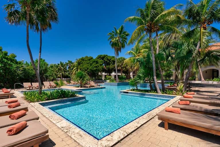 Tropical resort pool surrounded by palm trees, lounge chairs with orange towels, and clear blue sky