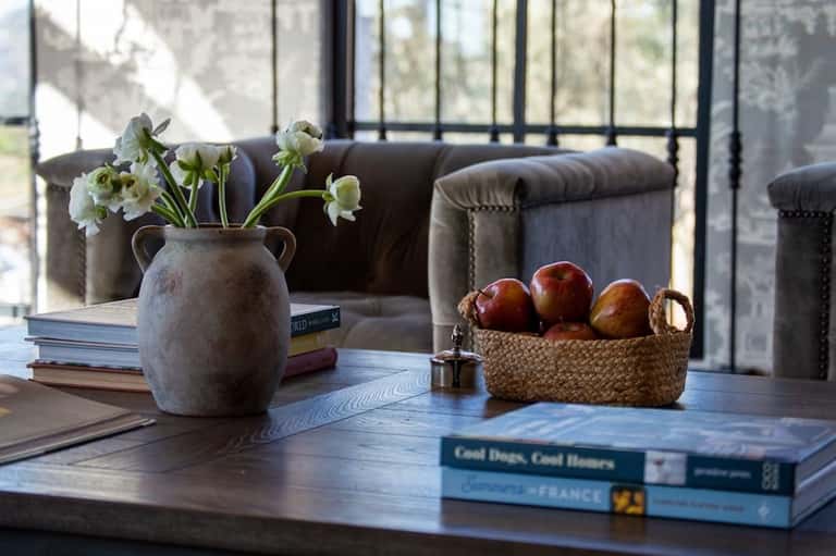 Wooden table with white flowers in ceramic vase, books, and fresh apples in woven basket