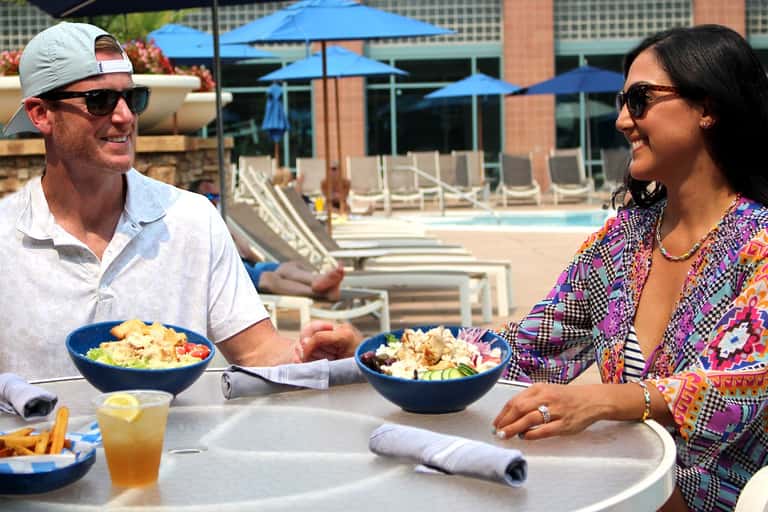 Two people dining poolside at a resort, with blue umbrellas and lounge chairs visible in the background