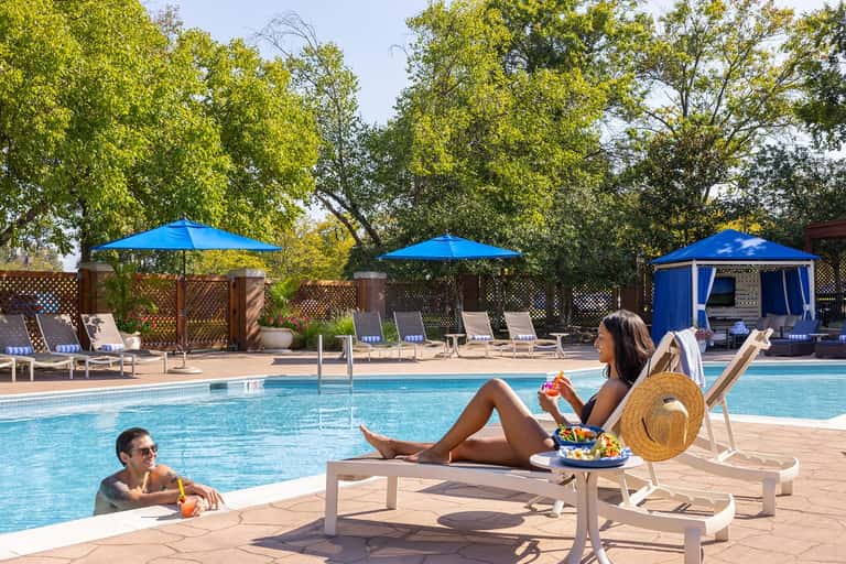 Resort pool area with swimmers and loungers, surrounded by trees and blue umbrellas for shade