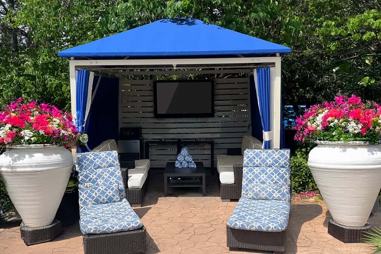 Outdoor gazebo with blue roof, TV, comfortable seating, and colorful flower planters on either side