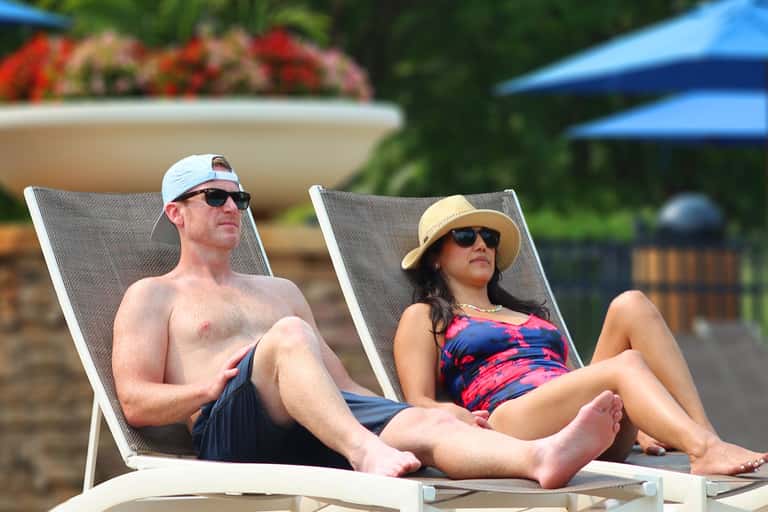 Couple sitting on lounge chairs by the pool, both wearing sunglasses and sun protection