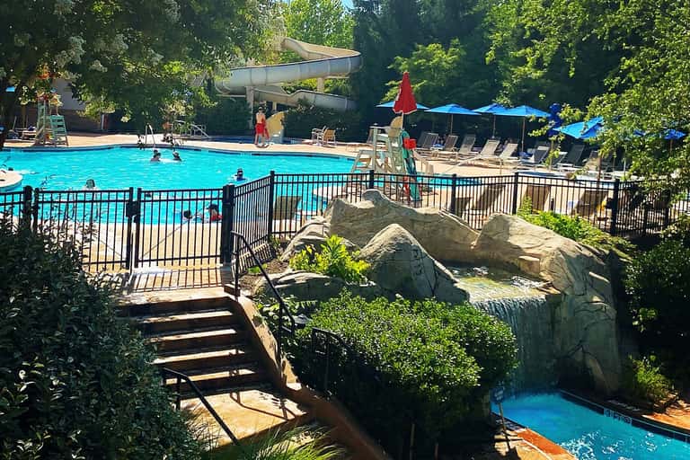 Large resort pool with waterslide, surrounded by trees, umbrellas, lounge chairs, and guests swimming