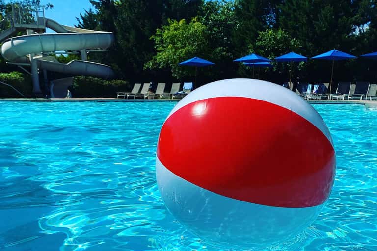 Red and white beach ball floating in a crystal-clear resort pool with waterslide in background