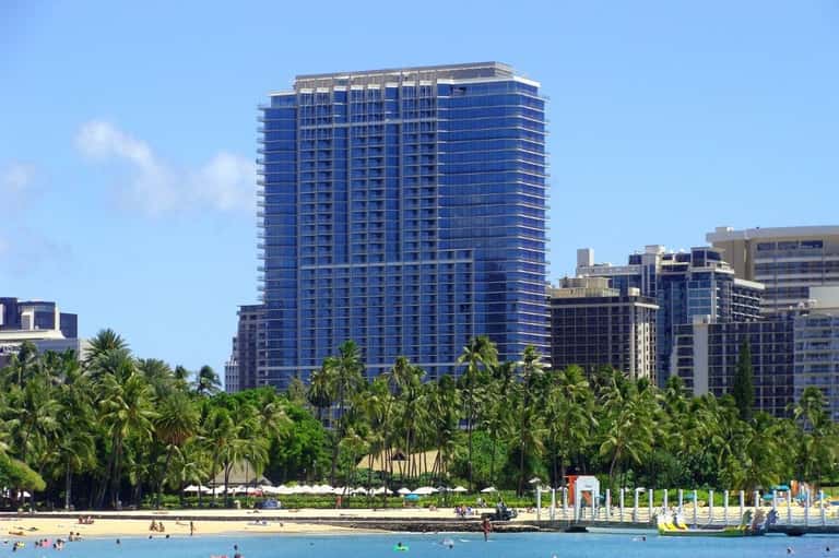Blue high-rise beachfront hotel with palm trees and sandy beach with swimmers in clear turquoise water