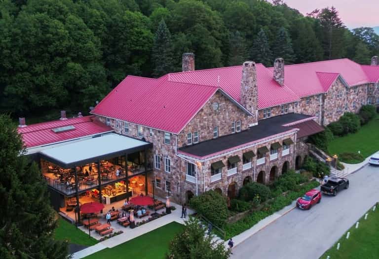 Stone luxury resort with pink metal roof, outdoor patio with red umbrellas, manicured grounds, and forest backdrop