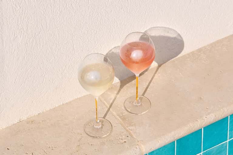 Two champagne glasses with white wine and rosé sitting on pool deck edge with shadows