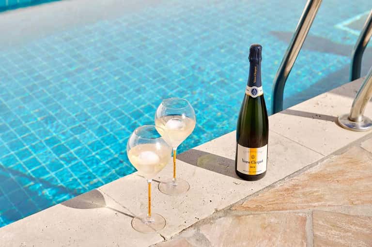 Two champagne glasses and bottle on pool deck edge with clear blue water in background