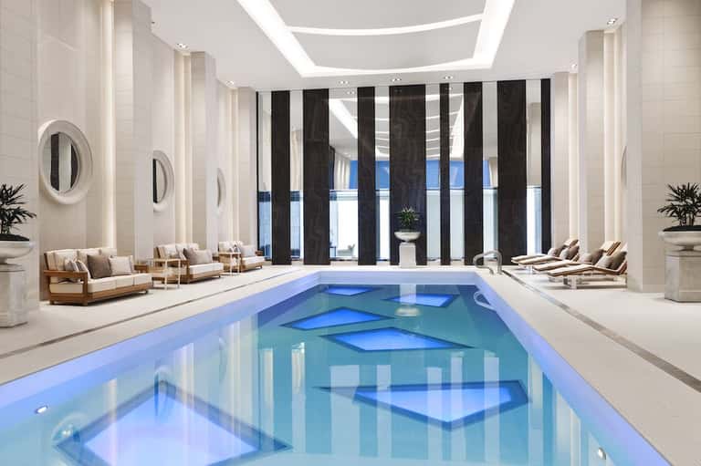 Luxury indoor swimming pool with blue lighting, elegant seating areas, and floor-to-ceiling windows with black striped curtains