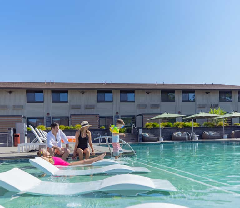 Family enjoying lounge chairs in a resort pool with modern building and umbrellas in background