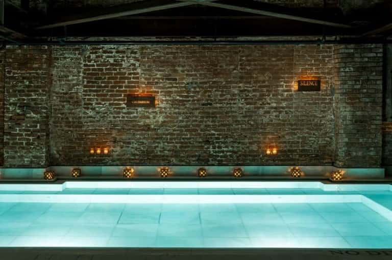 Indoor lap pool with illuminated turquoise water, weathered brick wall with 'CALDARIUM' and 'SILENCE' signs, golden lanterns